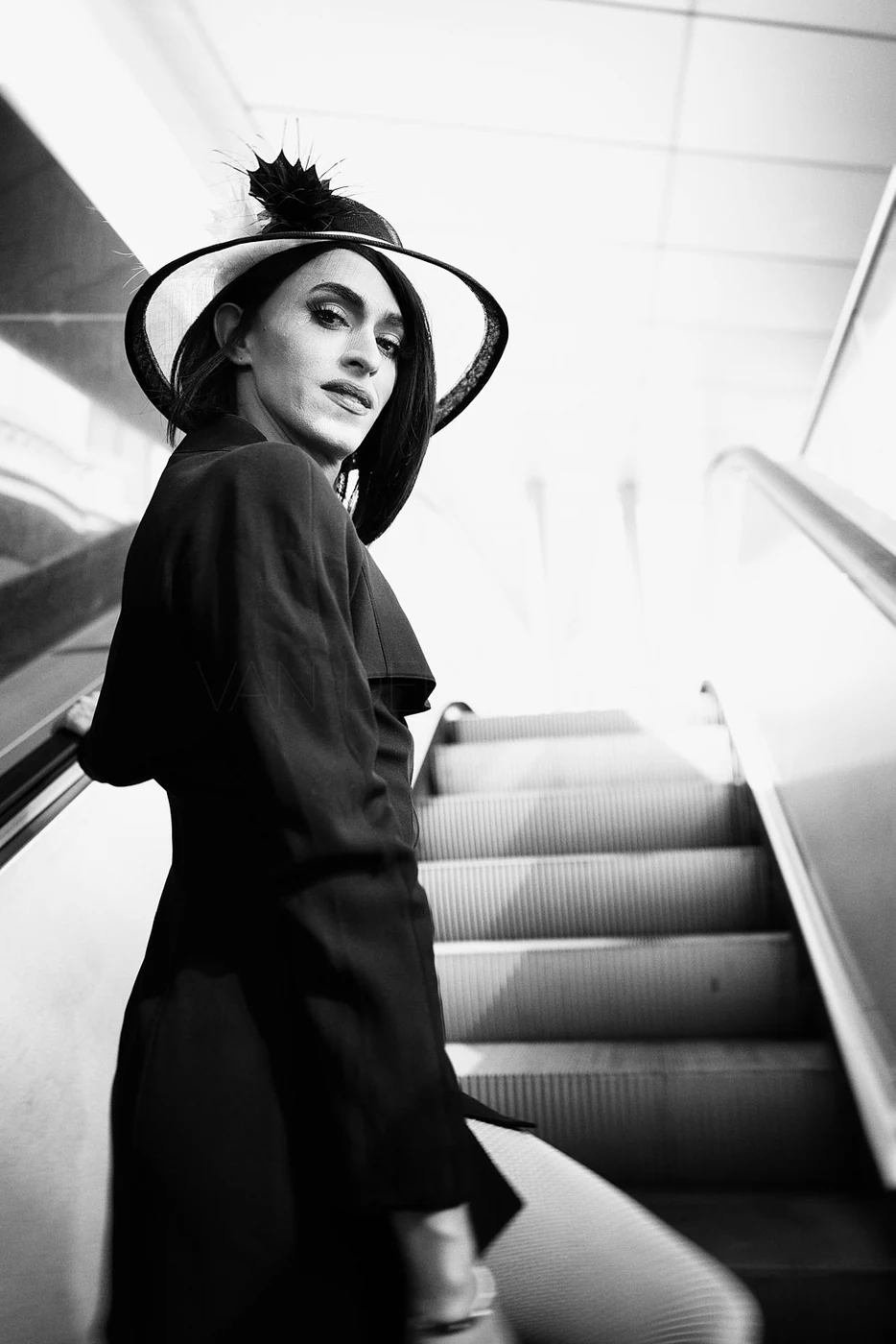 A woman wearing a large, ornate hat with a feather stands at the bottom of an escalator, looking back over her shoulder.