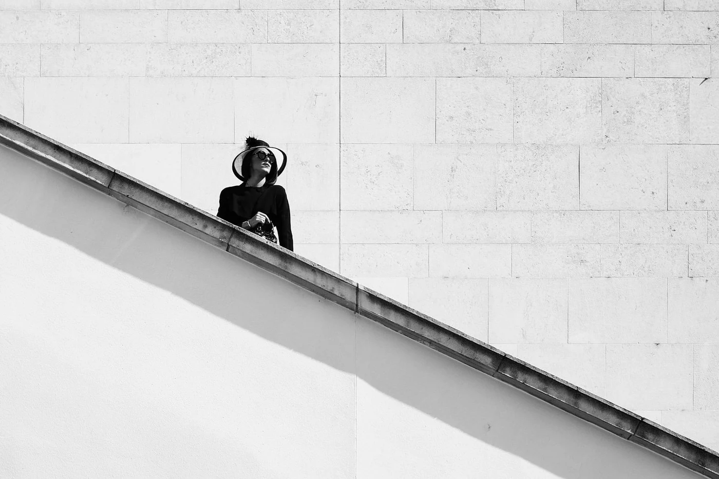 A woman wearing a hat, sunglasses, and dark clothing standing on an elevated balcony with a plain wall in the background, captured in black and white.