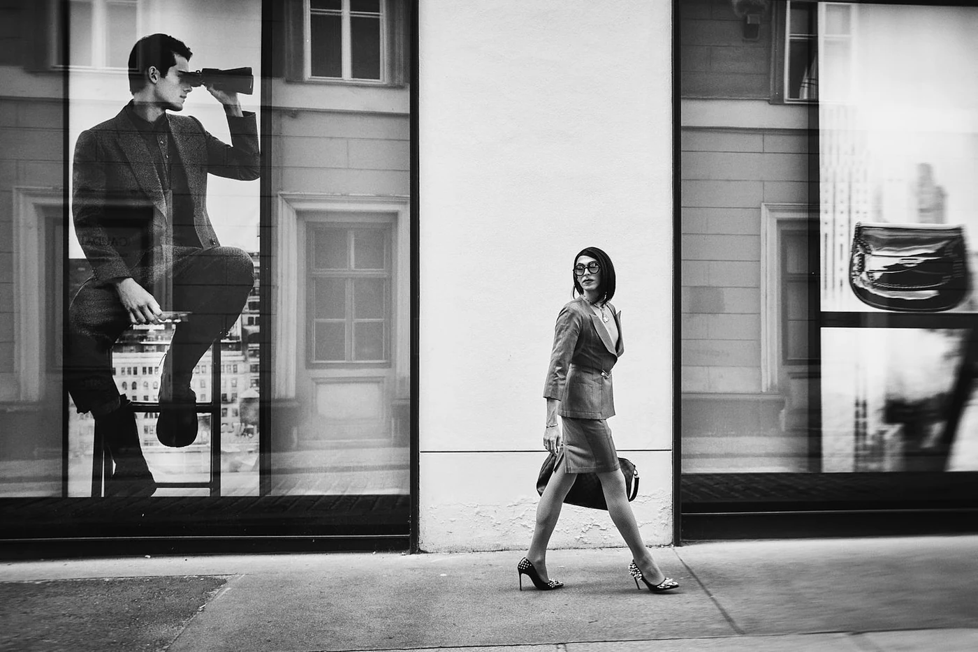 A woman in professional attire walking past store windows with large advertisements, one featuring a man in a suit looking through binoculars, in an urban setting.