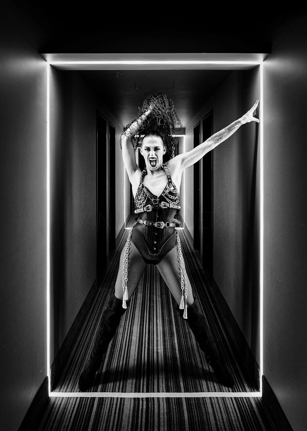 A fierce woman dressed in punk fashion, with chains and fishnet stockings, stands in a narrow corridor with her arms outstretched and mouth open in a roar, illuminated by bright LED lights framing the hallway.