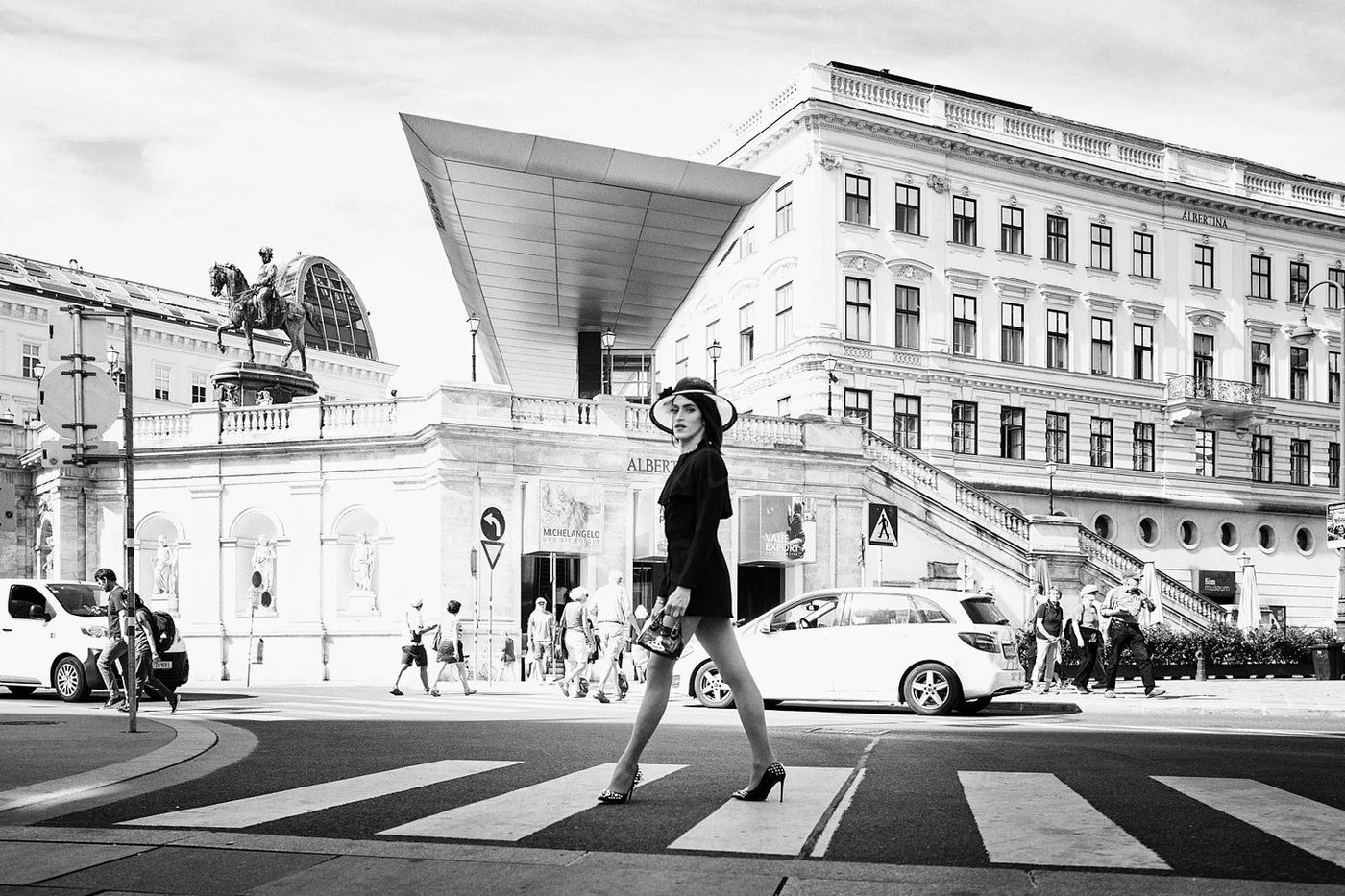 A woman in high heels and a dress crossing a city street zebra crossing, with historical and modern buildings and pedestrians in the background.