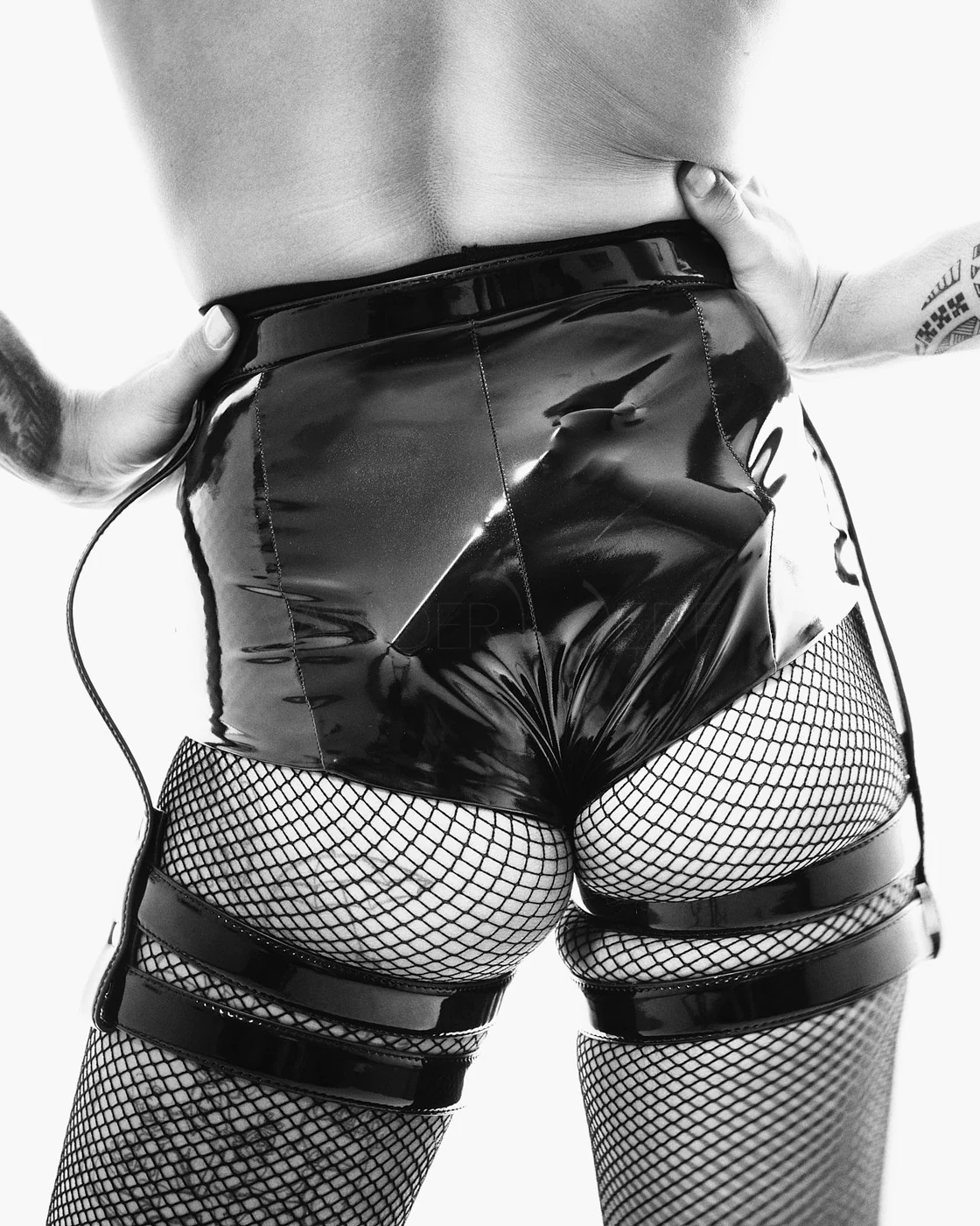 A person wearing shiny black shorts and fishnet stockings with black bands, standing with hands on hips.