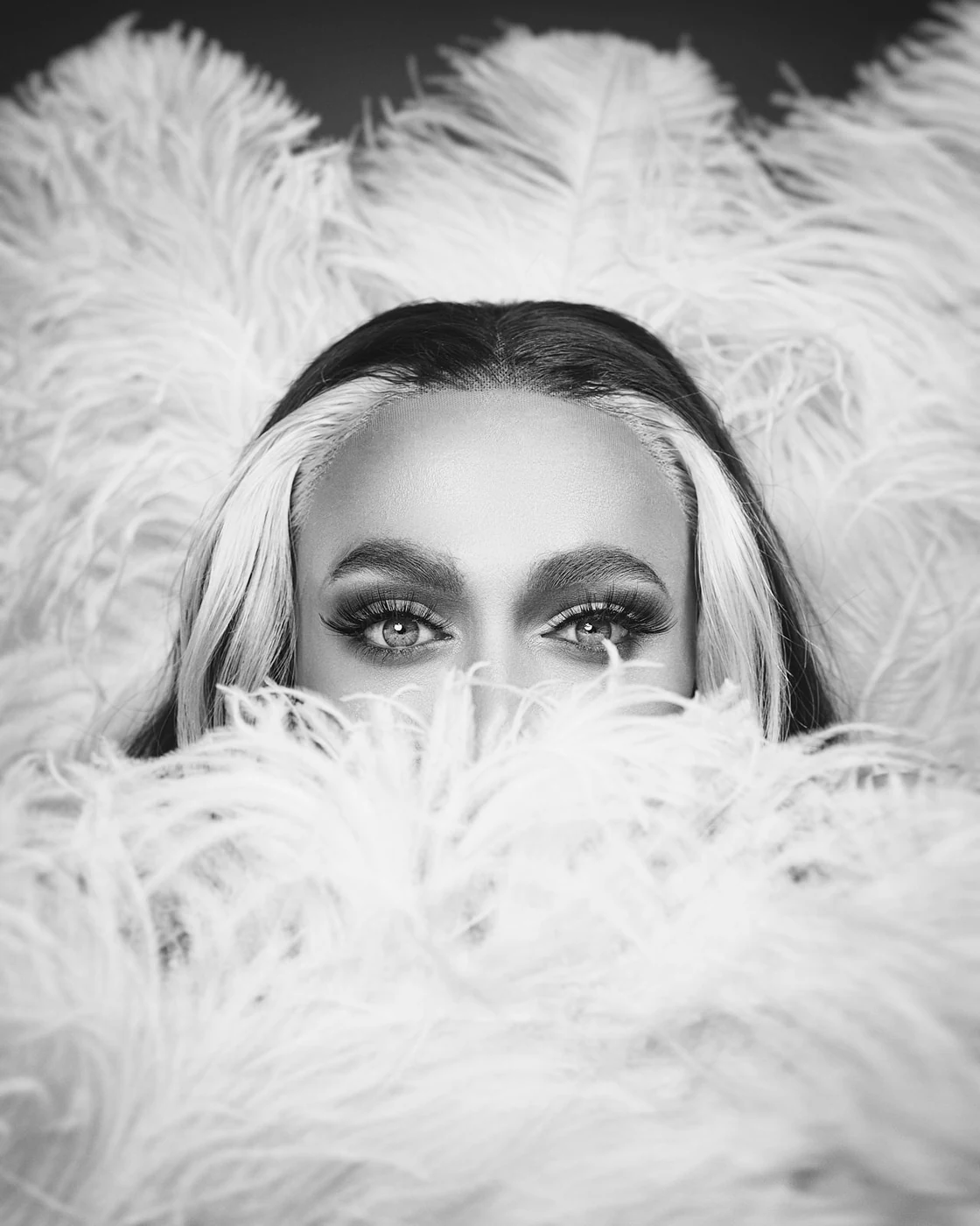 A black and white photo of a woman's face partially hidden behind fluffy feathers, with eyes looking directly at the camera.