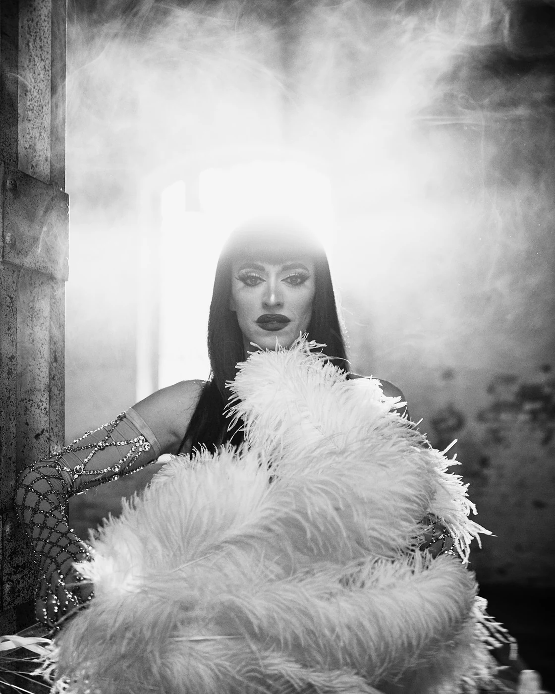 A woman with dark hair and makeup, wearing a feathered garment and jewelry, posing indoors with a bright light behind her in black and white.