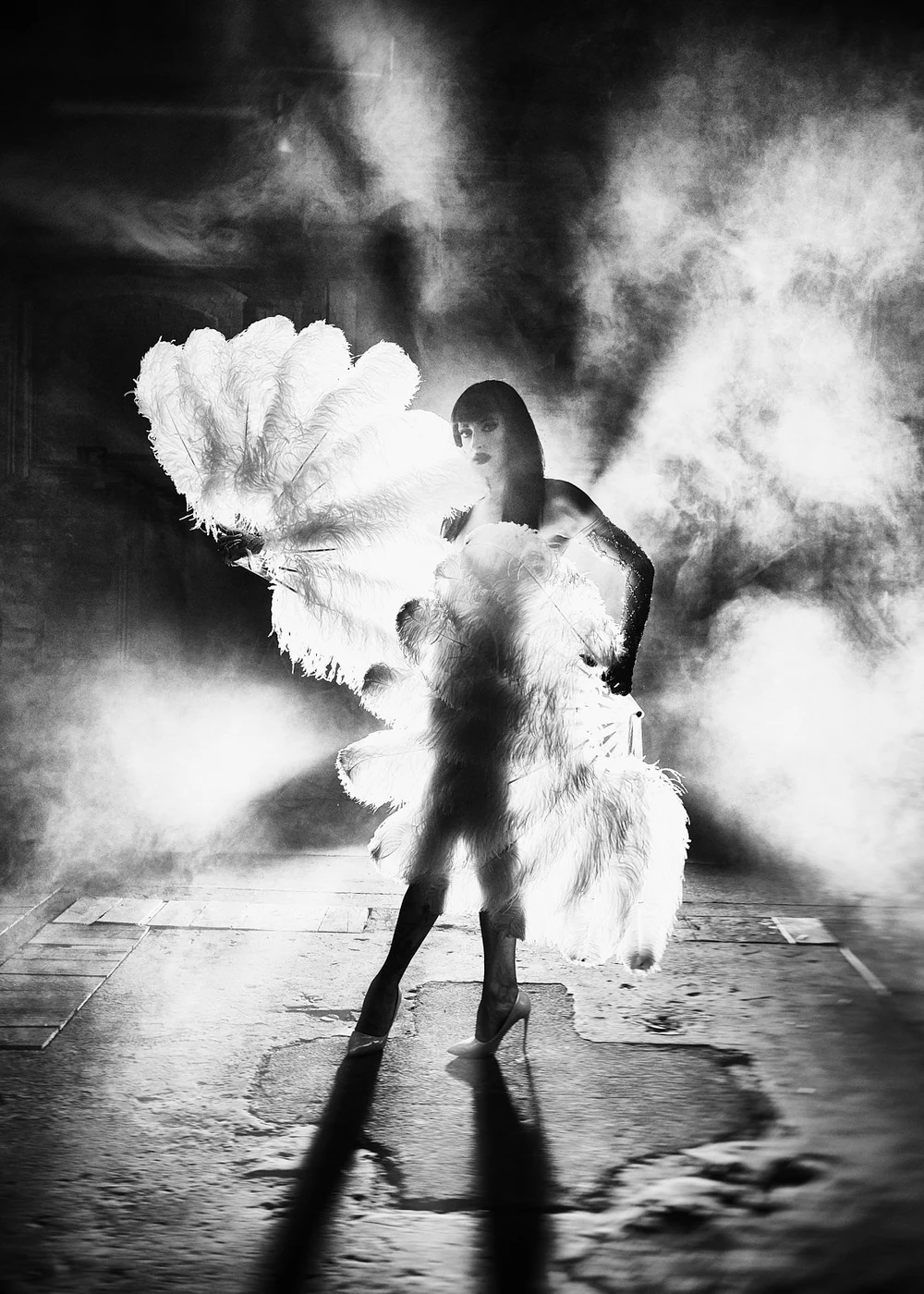 A woman in high heels and dark clothing stands on a street at night, surrounded by smoke or fog, holding large white feather fans.