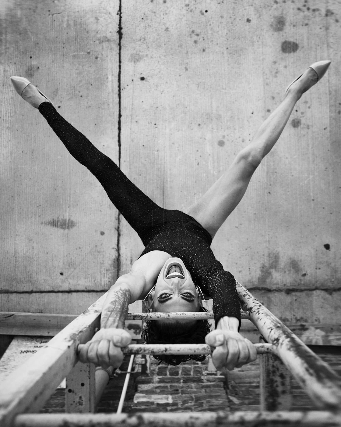 Black and white photo of a woman in a sparkly black leotard performing a contortion on a metal ladder, with her head upside down and mouth open, against a concrete wall background.