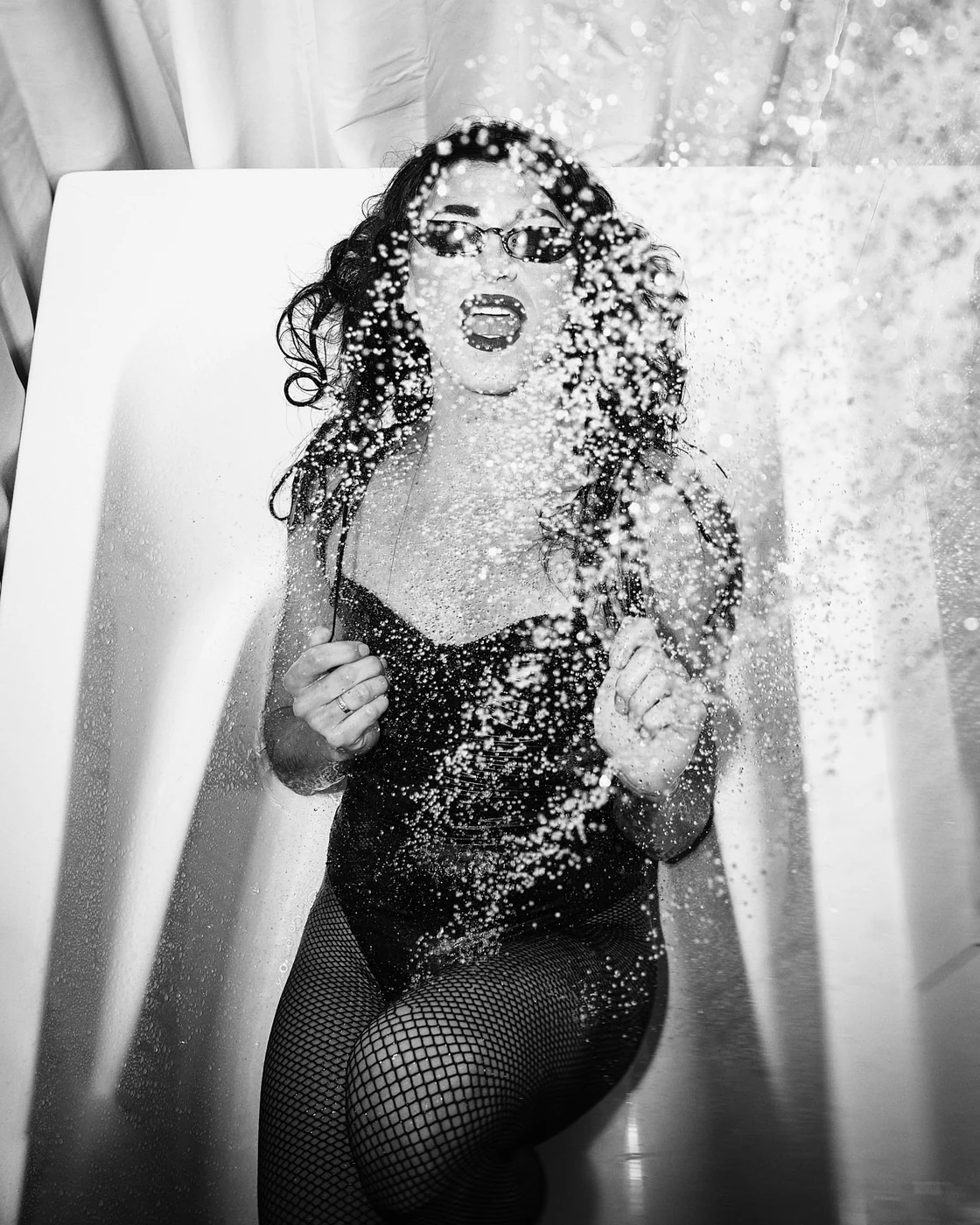 A woman with glasses and curly hair wearing fishnet stockings and a sleeveless top is behind a glass shower door, with water splashing onto the camera lens, creating a dynamic and artistic scene.