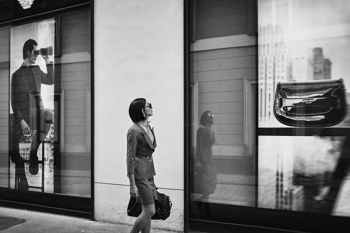 A woman with sunglasses, dressed in business attire, standing outside a modern building with large windows. Reflections of other people and cityscape are visible in the windows, including a man taking a photo with a camera.