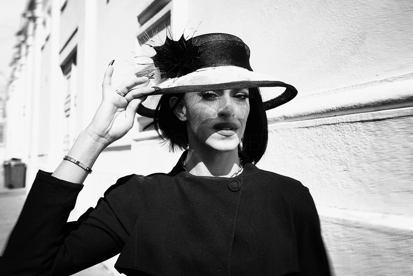 A woman wearing a large hat with feather decoration, holding the brim with her right hand, standing outdoors near a textured wall, in black and white.