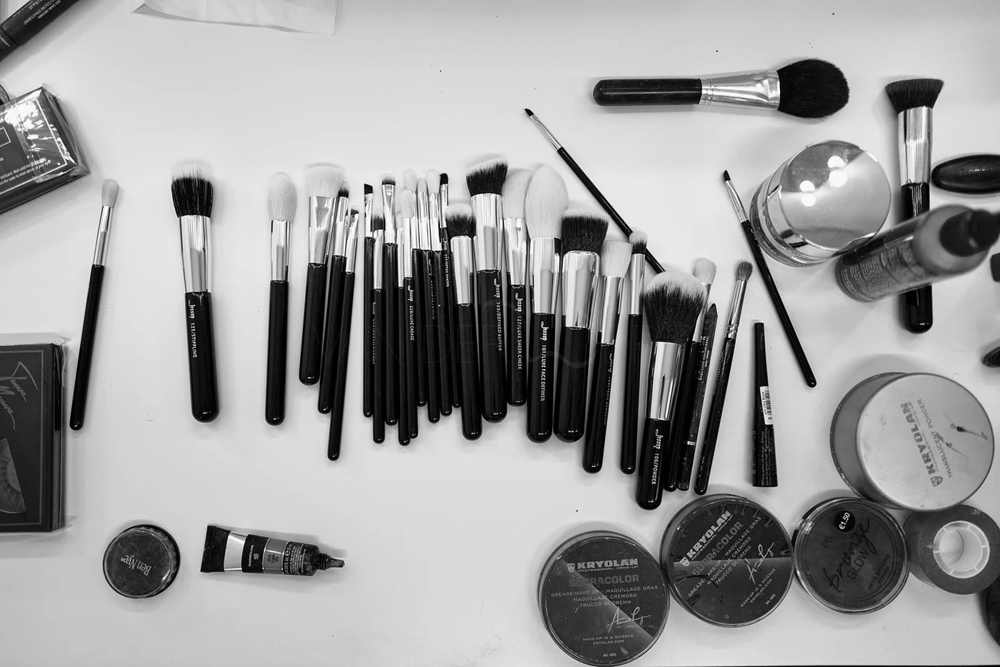 Beauty makeup brushes, powders, and cosmetic products arranged on a white surface, including various brushes, a small jar, and containers of makeup.