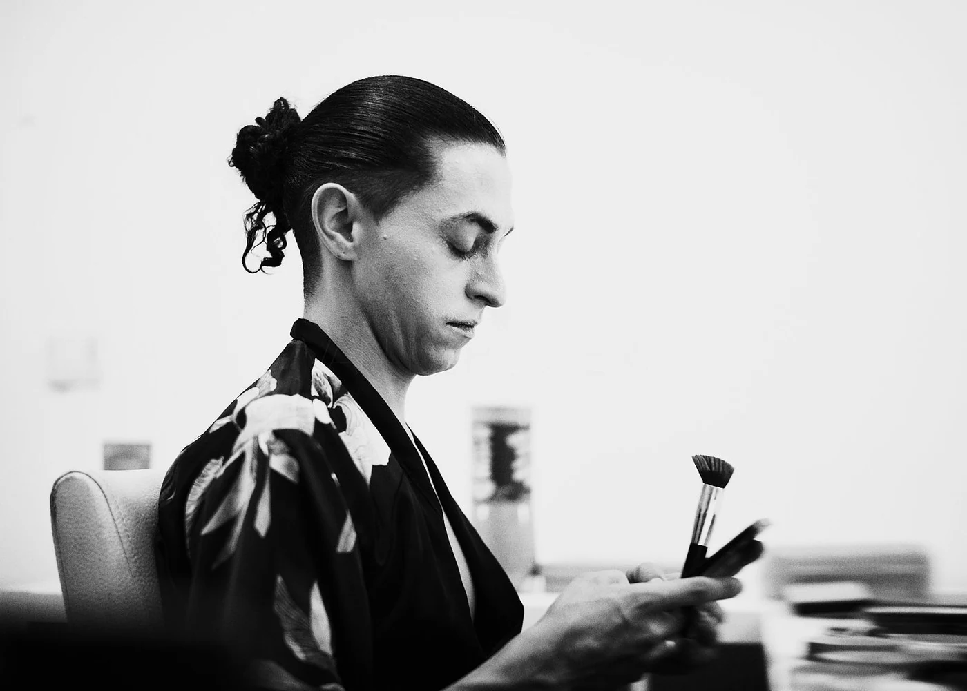 Black and white photo of a woman sitting at a desk, looking at her phone with a makeup brush in hand