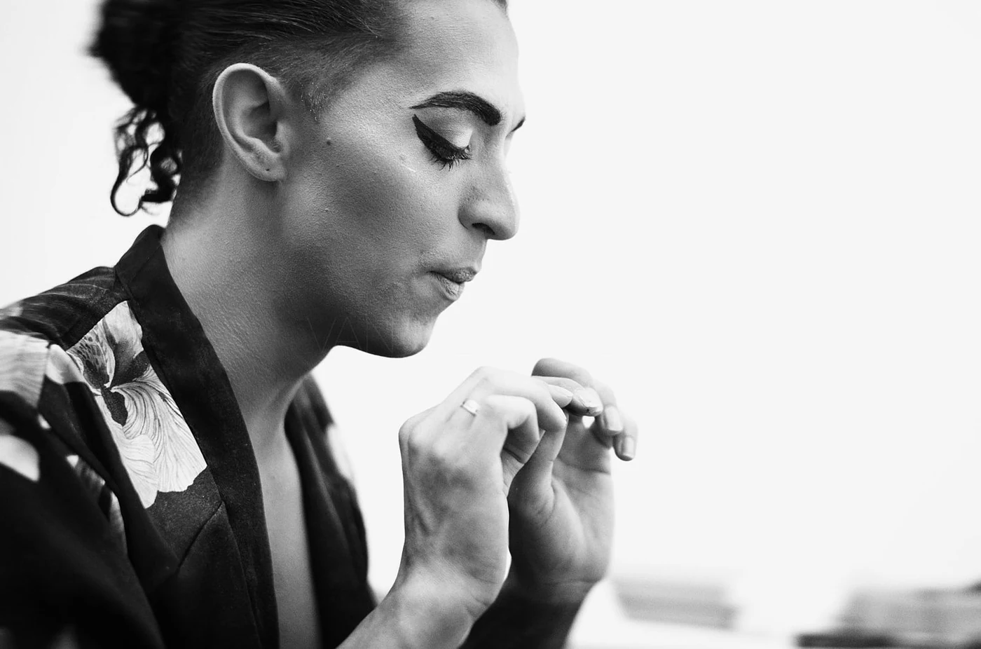 Black and white close-up of a woman with dark hair styled in an updo, wearing eyeliner and a patterned jacket, looking down with her hands close to her face.