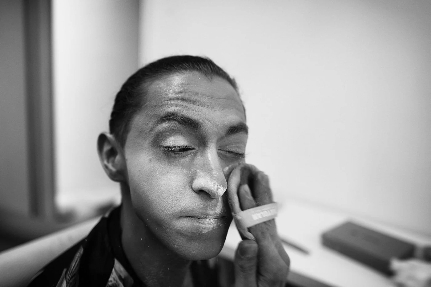 A person with closed eyes getting their makeup applied by a hand wearing a band on the finger in black and white.