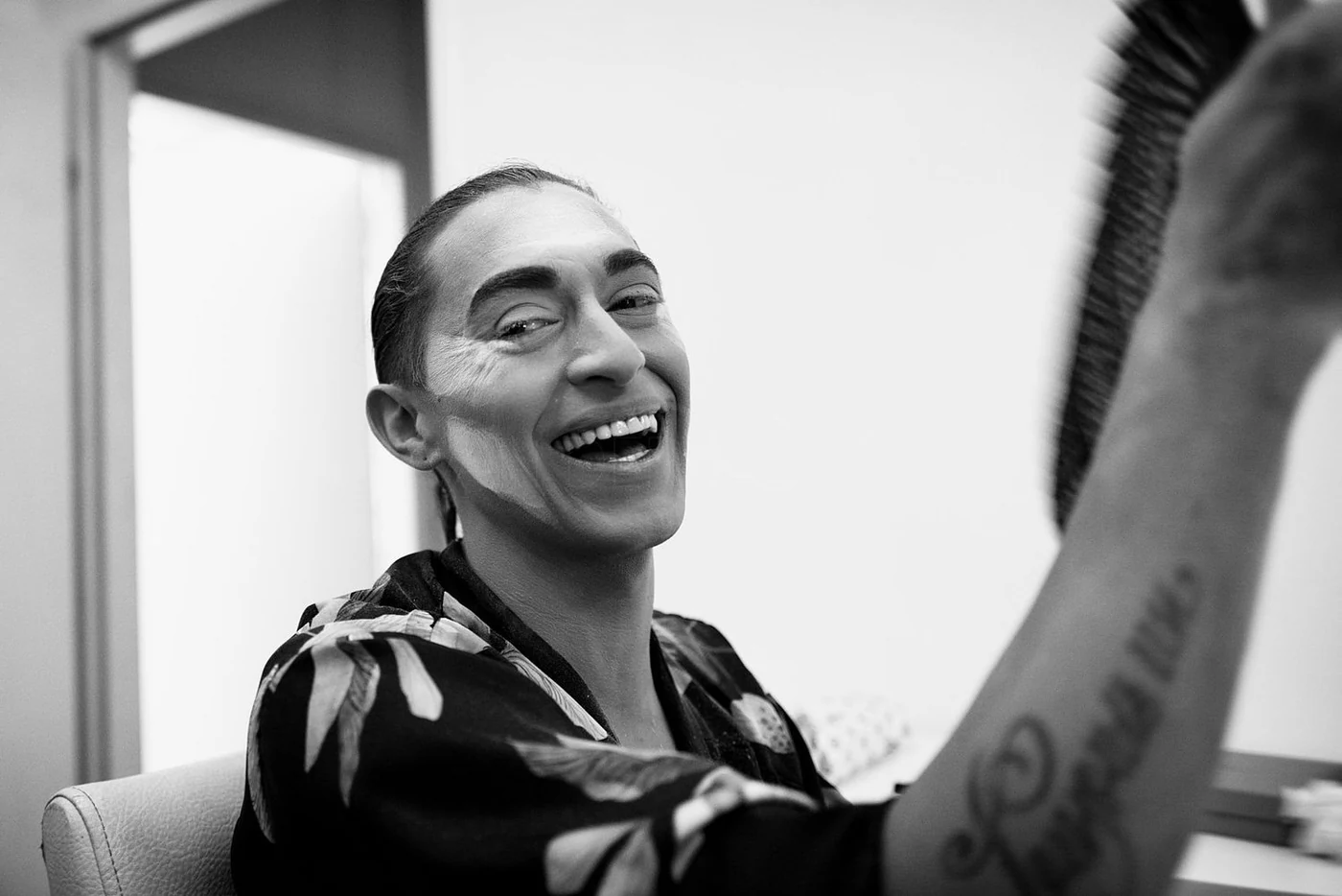Black and white photo of a smiling woman with short hair, wearing a patterned shirt, sitting in a chair, with part of her arm and face visible in the foreground.