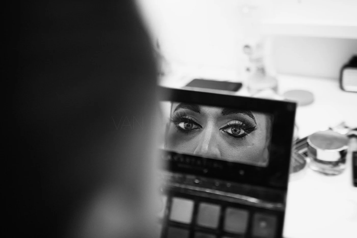 Close-up of a woman's eyes reflected in a makeup mirror, with makeup products and tools on a desk in the background, in black and white.