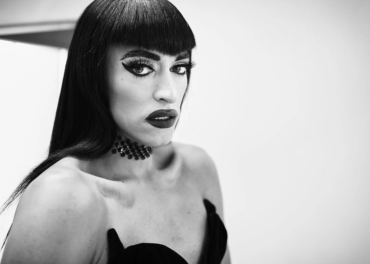 A woman with dark hair and dramatic makeup, wearing a strapless black dress and choker, looking at the camera against a plain background.