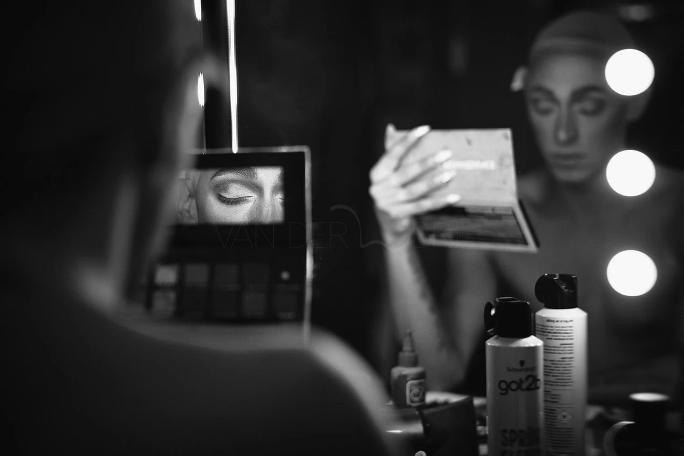 A woman with her eyes closed applying makeup in front of a mirror with bright round lights. The mirror reflects her face, and she is holding a makeup palette or book. There are various makeup and skincare products on the table.