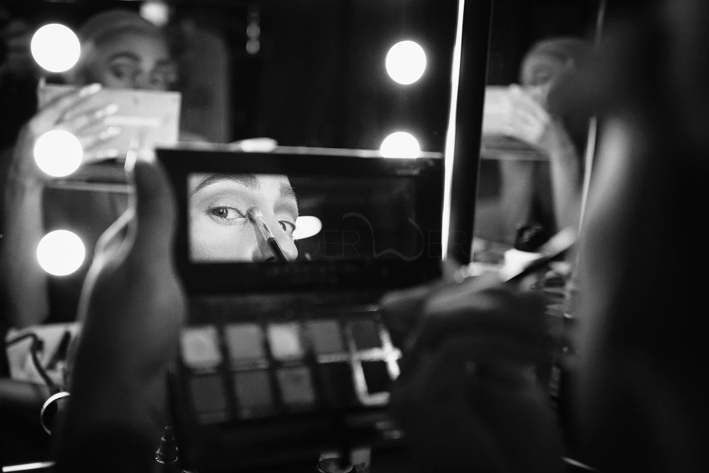 A woman applying eye makeup while looking into a mirror, with her reflection visible in a makeup mirror surrounded by bright round lights. The photo is in black and white.