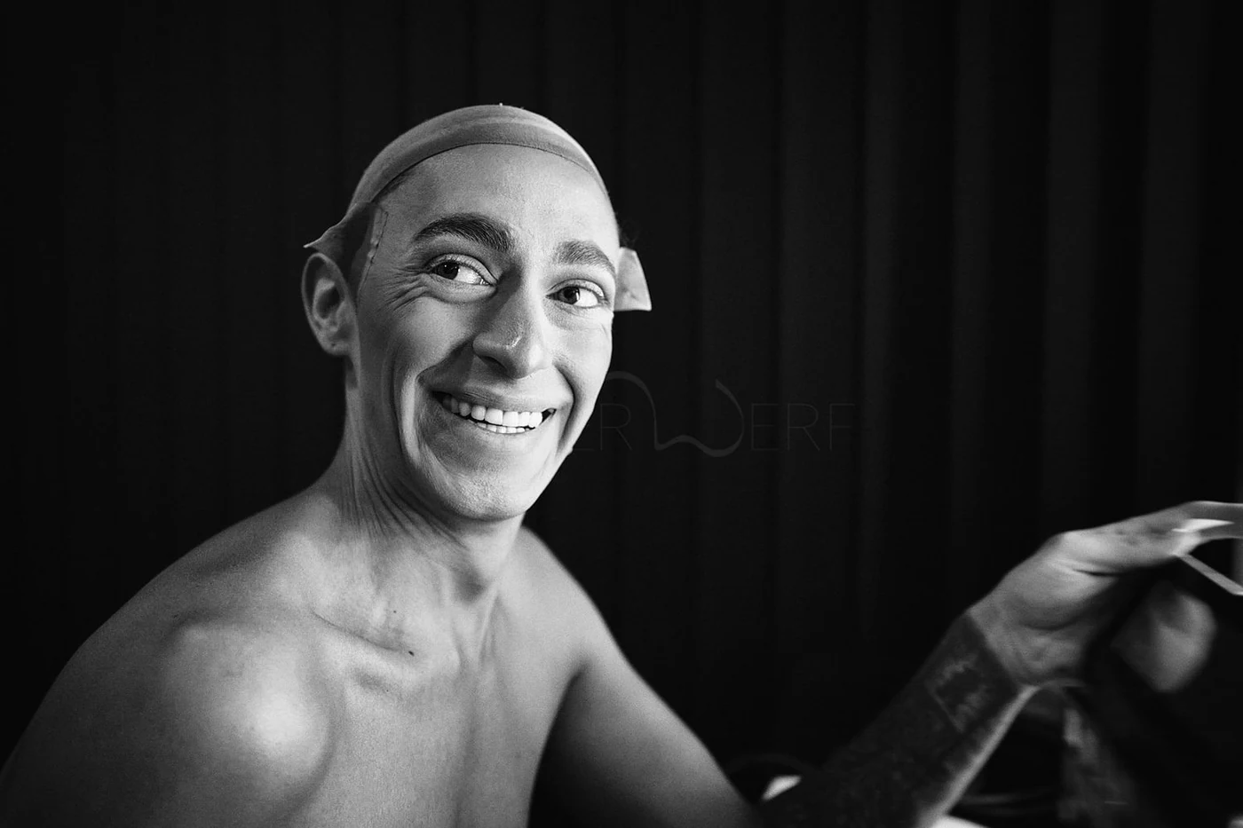 Black and white photo of a smiling man without a shirt, wearing a cap backwards and looking to the left, with a tattoo on his left arm