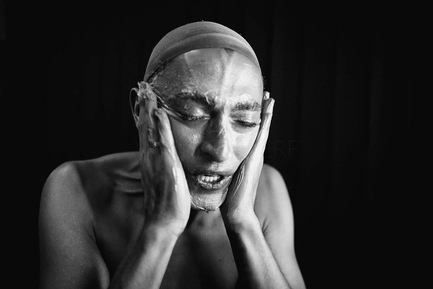 A black and white photograph of a woman with her eyes closed, washing her face with water, with water dripping down her face, in front of a dark background.