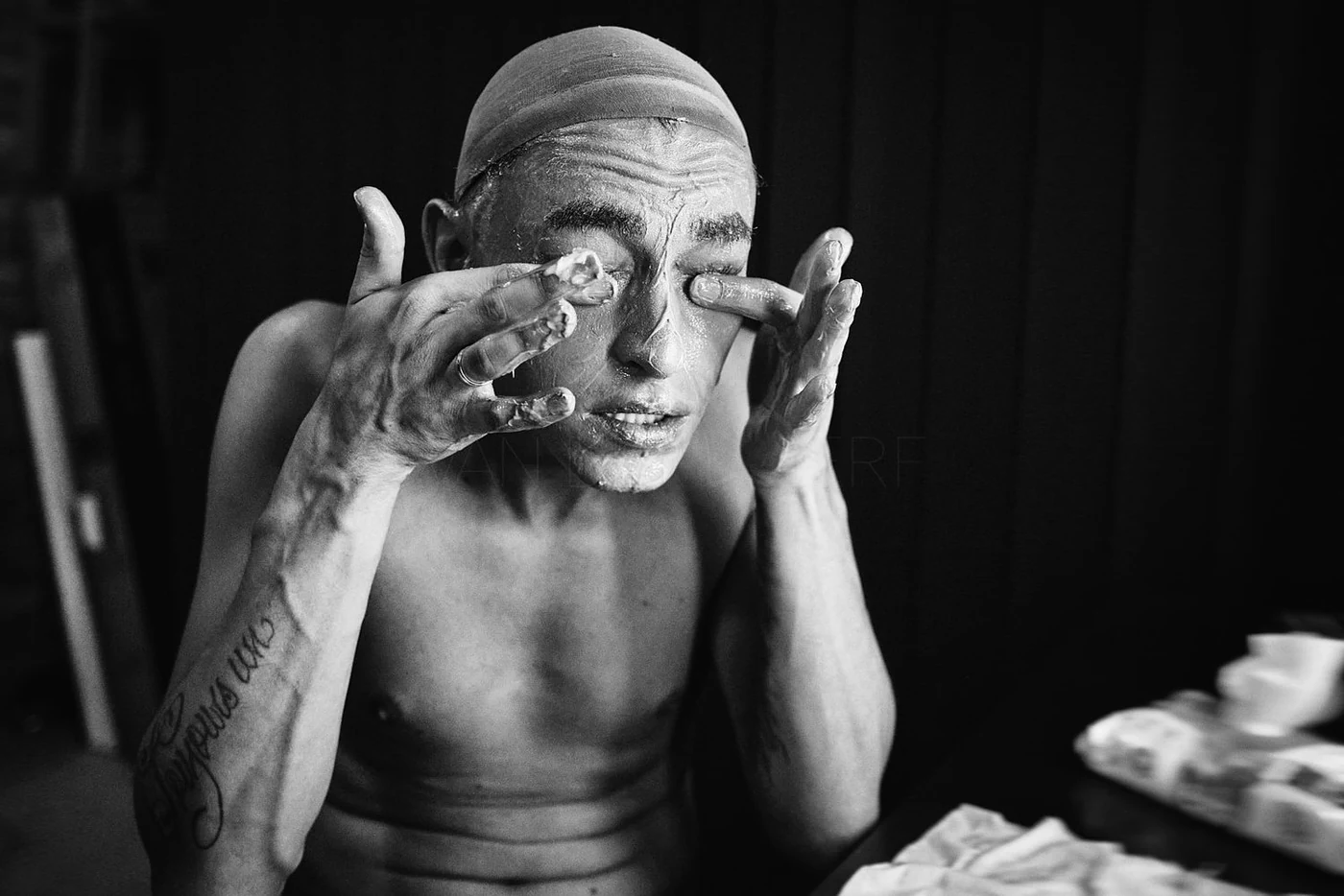 A shirtless person with tattoos, wearing a cap, applying soap or cream to their face, in black and white.