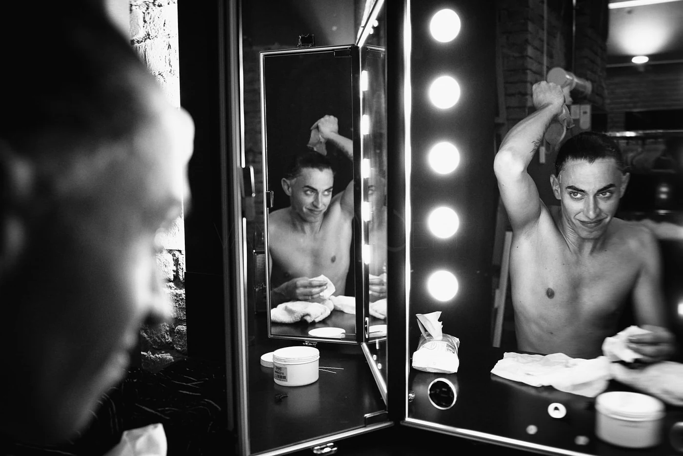 A shirtless man with dark hair is in front of a mirror, looking at his reflection while holding a towel and a spray bottle. The setting appears to be a backstage or dressing room with bright lights around the mirror. The scene is in black and white.