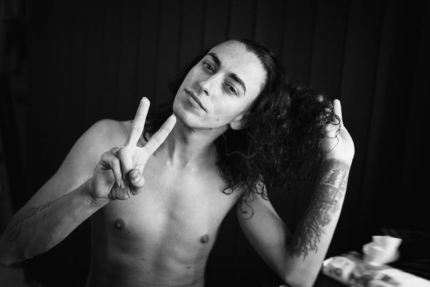Black and white photo of a shirtless man with long curly hair making a peace sign and tilting his head.