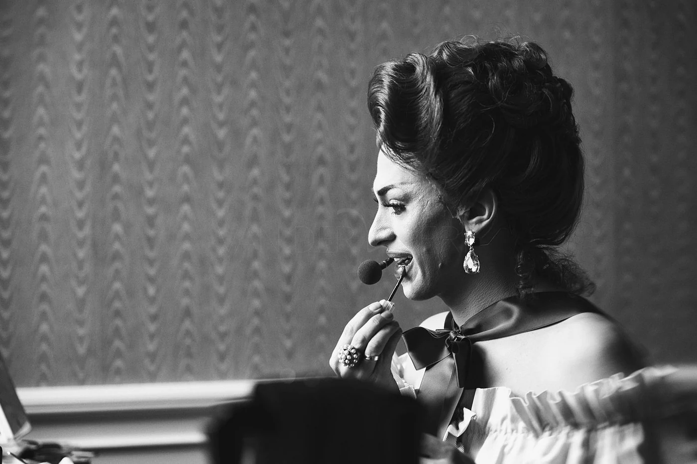 A black and white photo of a woman with styled hair and large earrings applying lipstick with a brush.