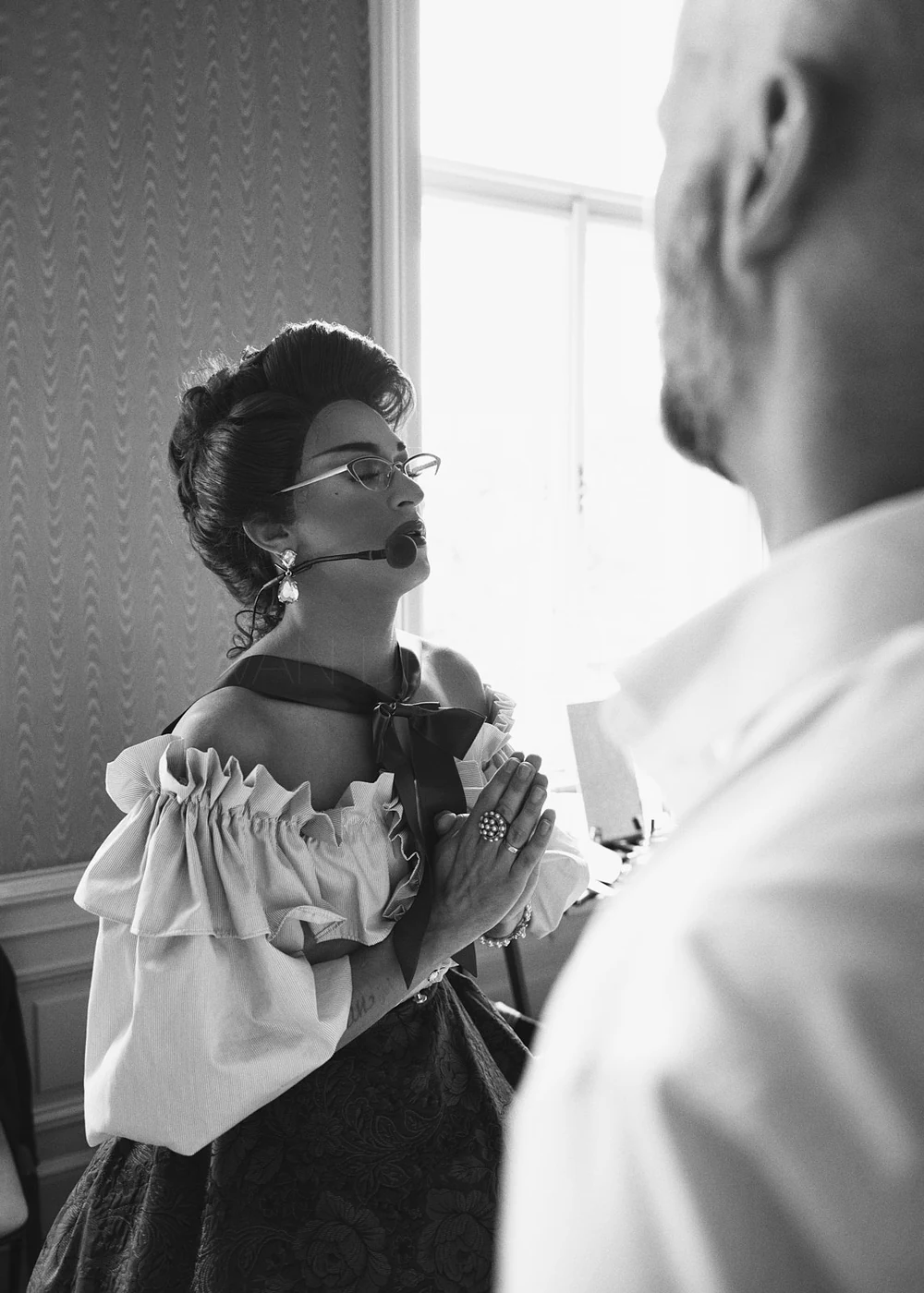 A woman with styled hair, glasses, and earrings praying with her hands pressed together, wearing a ruffled off-the-shoulder top and a bow tie, standing in profile inside a room with a window, with a man partially visible in the foreground.