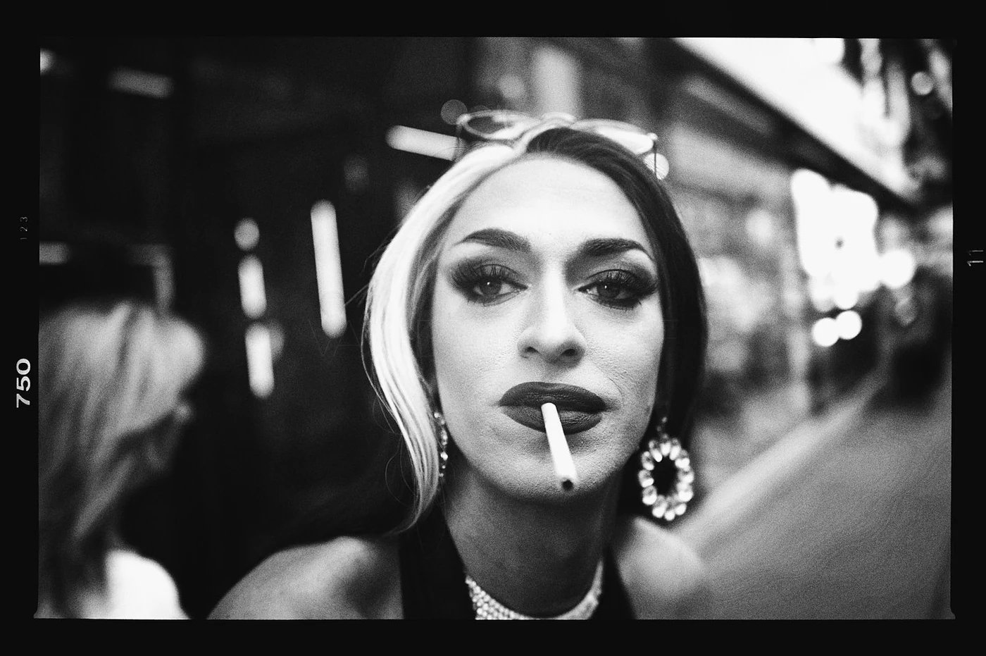 Close-up black and white photo of a woman with striking makeup, dark lipstick, and earrings, holding a cigarette in her mouth, on a busy street.