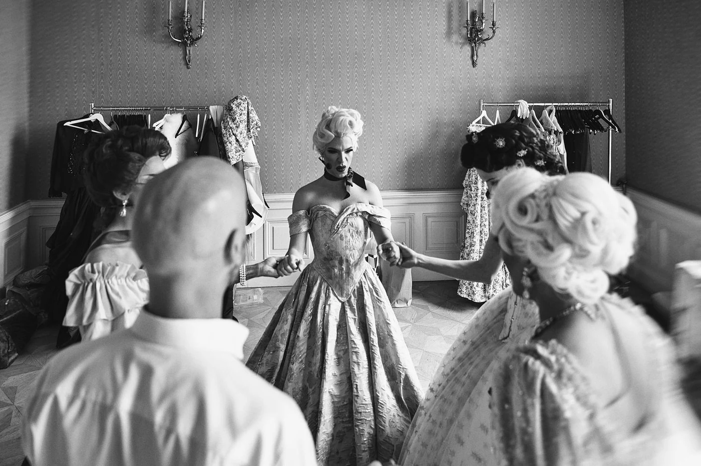 A black and white photo of a woman dressed in a vintage, elaborate gown holding hands with three other women in a dressing room, with clothing racks in the background.
