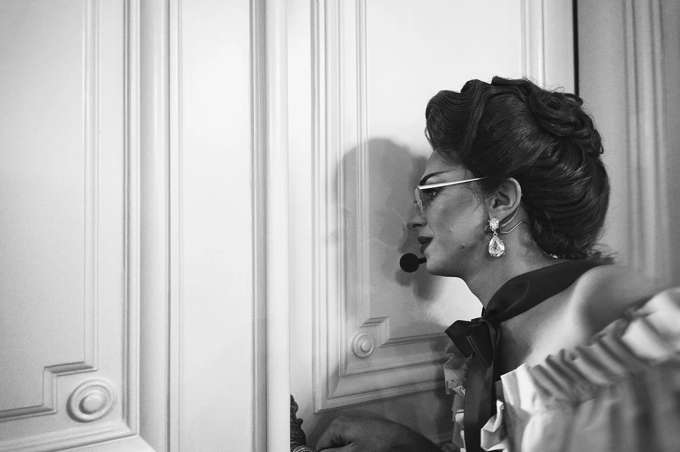 A woman with styled hair, wearing glasses, earrings, a bouquet, and a microphone headset, standing against a detailed paneled wall.