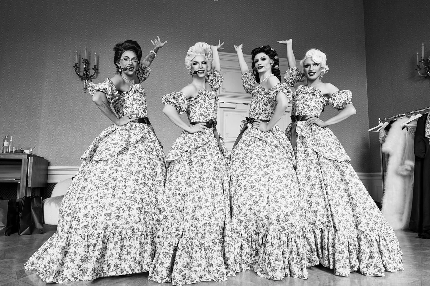 Four women dressed in vintage floral gowns with puffed sleeves and full skirts, posing confidently in a room with patterned wallpaper and elegant sconces.