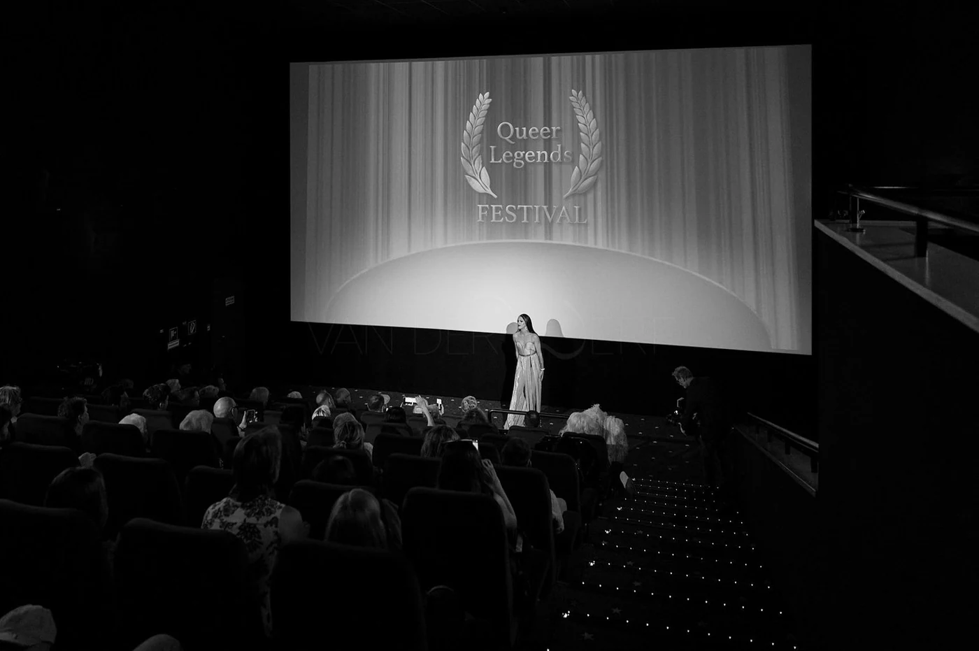 A woman stands on stage at the Queer Legends Festival, with a large screen behind her displaying the festival's logo, a laurel wreath and the words 'Queer Legends FESTIVAL'. The audience is seated in dark auditorium.