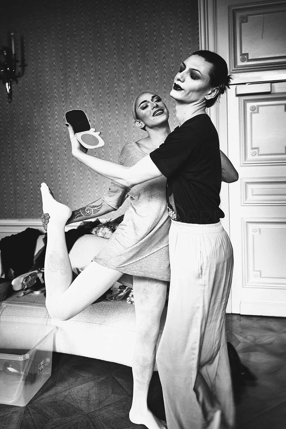 Two women taking a selfie together in a dressing room, one holding a makeup mirror and the other making a funny face. One woman has makeup on and is lifting her leg, showing tattoos on her thigh. The image is in black and white.