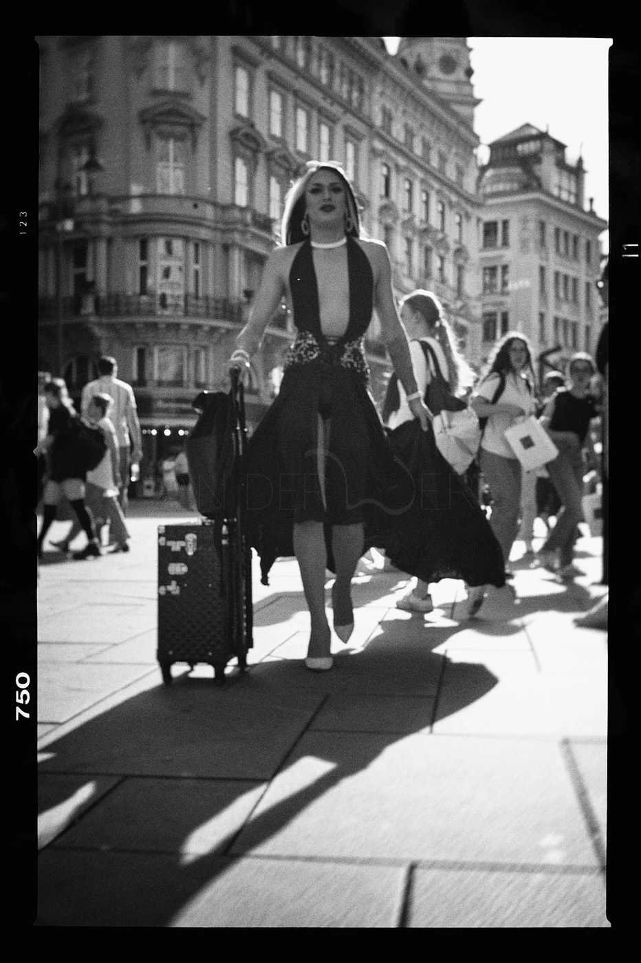 A woman in fashionable clothing pulling a wheeled suitcase walks through a busy city street with large buildings in the background.