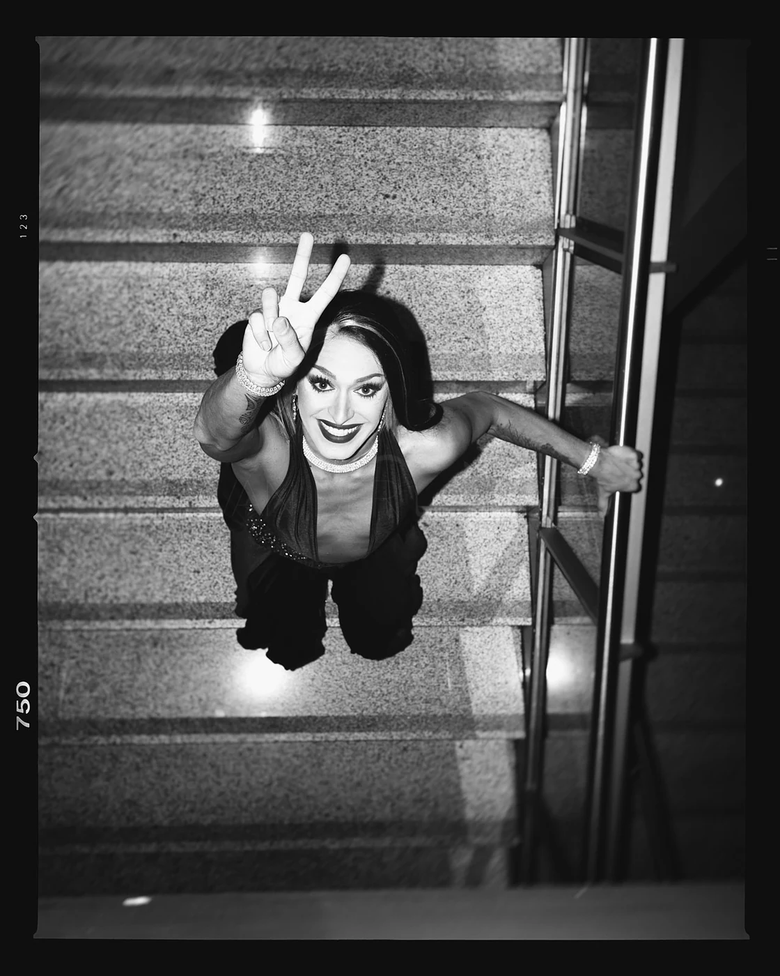 A woman with dark hair smiling and making a peace sign, looking up at the camera from a staircase, taken from above in black and white.