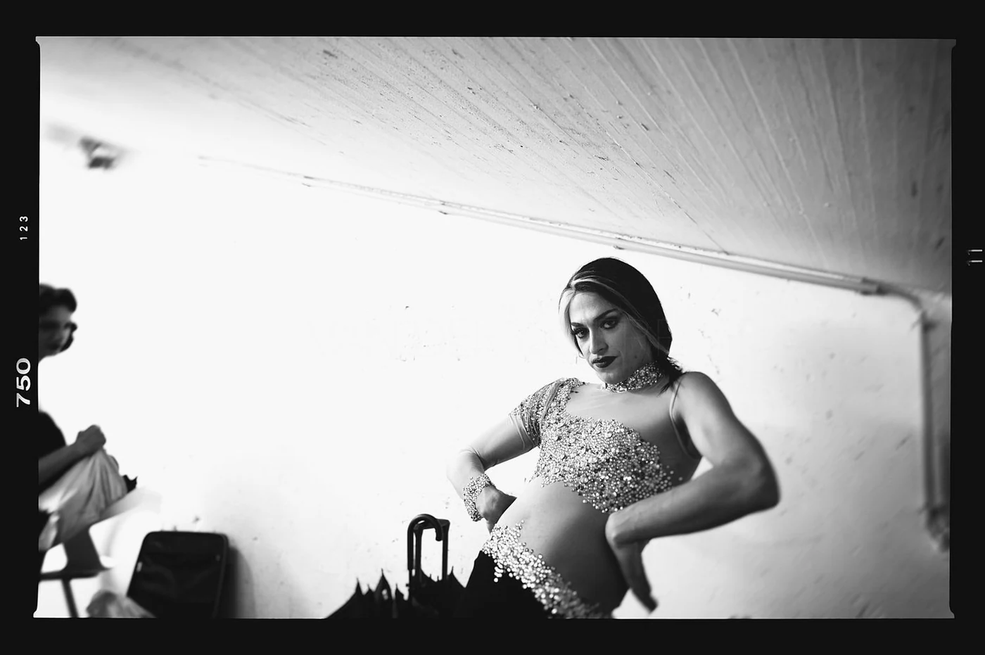 Black and white photo of a woman in a fitted, bejeweled top with her hands on her hips, looking confidently at the camera in a room with a plain wall and ceiling.