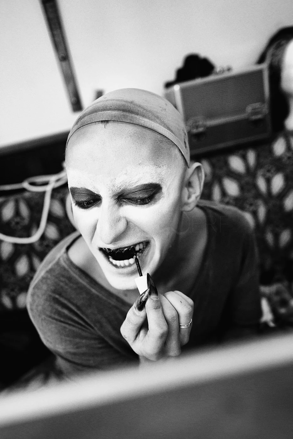 A person with makeup and a head covering applying lipstick while smiling and appearing to have vampire-style teeth, in a black-and-white photo.