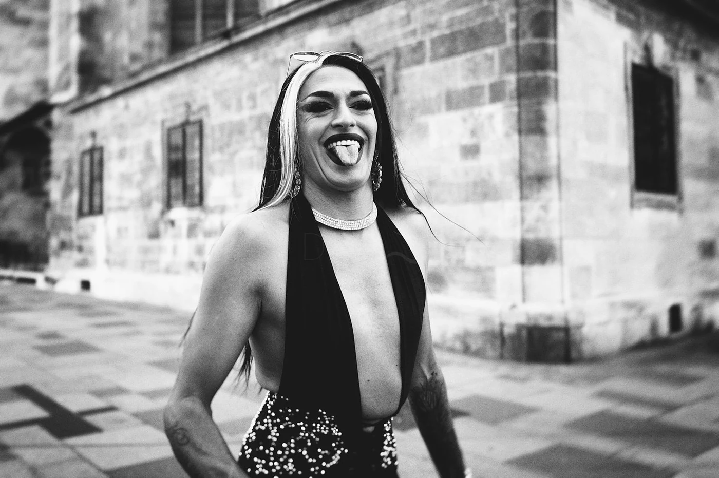 Black and white photo of a woman with long hair, wearing a deep V-neck dress with sequins, smiling and sticking out her tongue, standing outdoors in front of a stone building.
