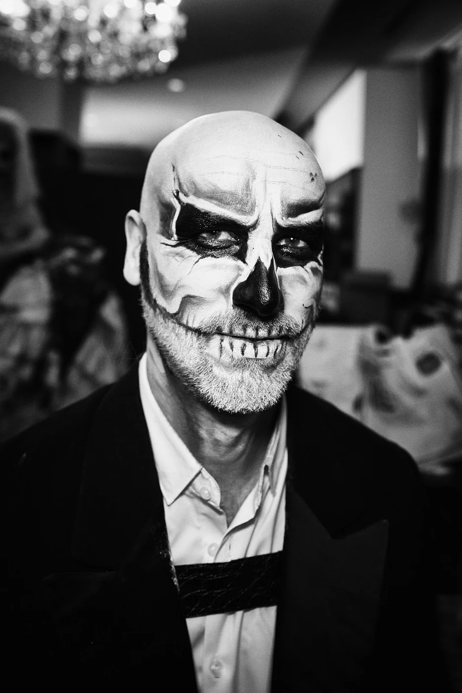 Man with skull and skeleton face paint, dressed in a tuxedo, with a white beard, at a celebration or costume event.