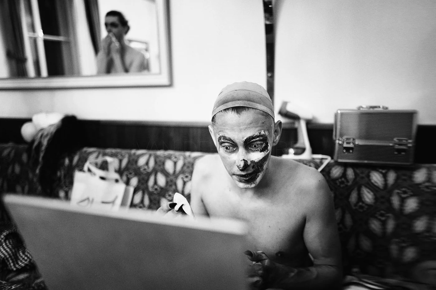 A person with makeup resembling a clown, wearing a head covering, is seated shirtless on a couch in a room, looking at a laptop. Another person is visible in the background looking into a mirror.