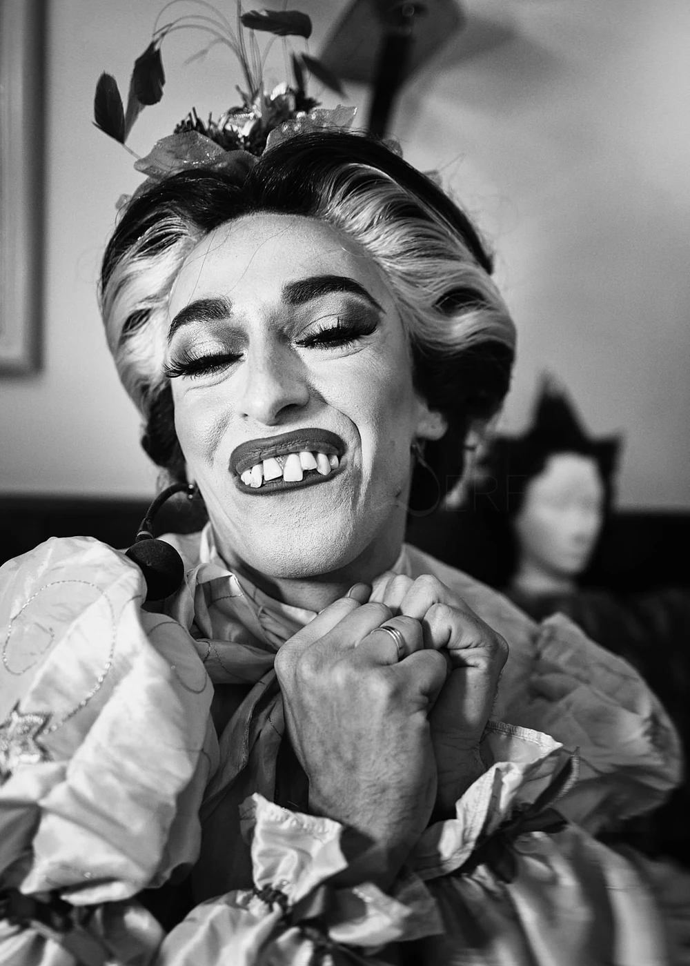 Black and white photo of a woman dressed as a clown with exaggerated makeup, large teeth, and a playful expression, clasping her hands together under her chin, with a blurred figure in the background.