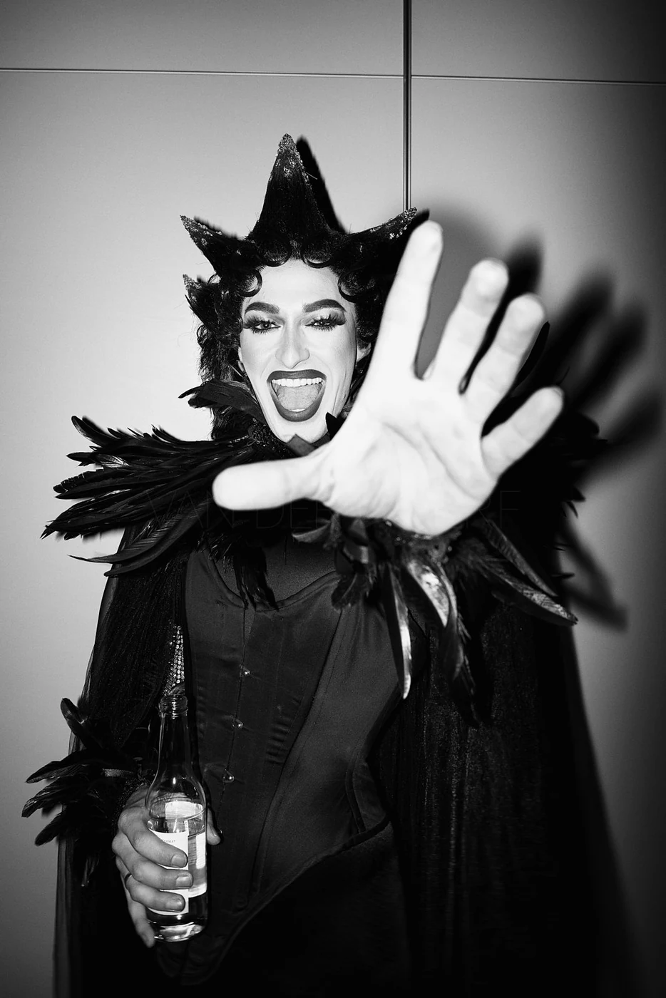 A person in a dramatic black costume with feathered details, making a gesture towards the camera with an open mouth and wide eyes, holding a bottle in one hand.