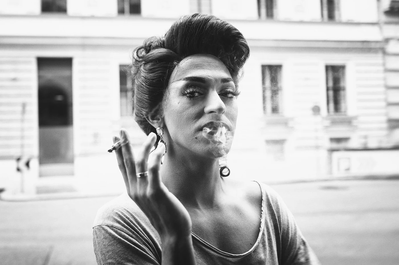 Black-and-white photo of a woman with styled hair smoking a cigarette outdoors, wearing earrings and a casual top, with a blurred urban building in the background.