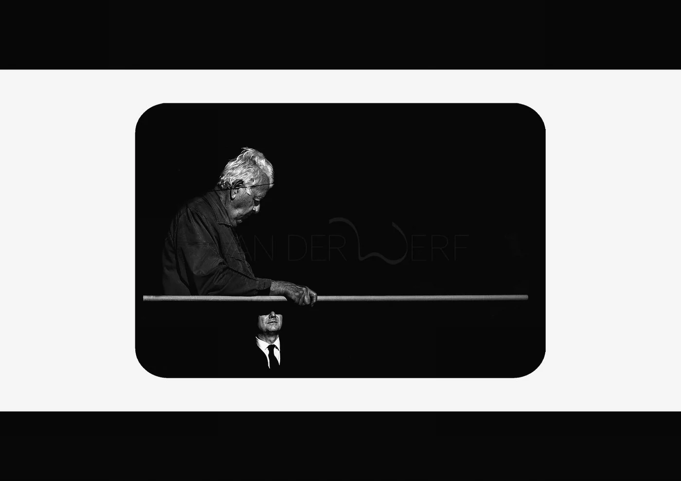 Black and white image of an elderly man with white hair looking down, holding a long stick, with a reflective surface below showing a man in a suit and tie.