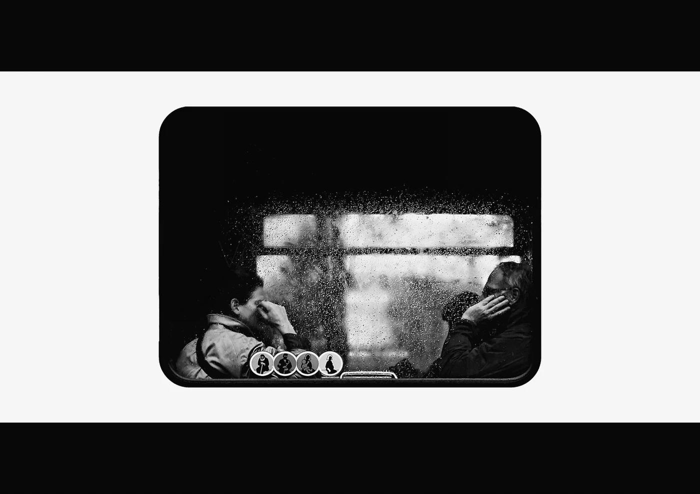 Black-and-white photo of two people sitting on a bus or train, viewed through a rain-covered window, appearing to be in distress or deep thought.