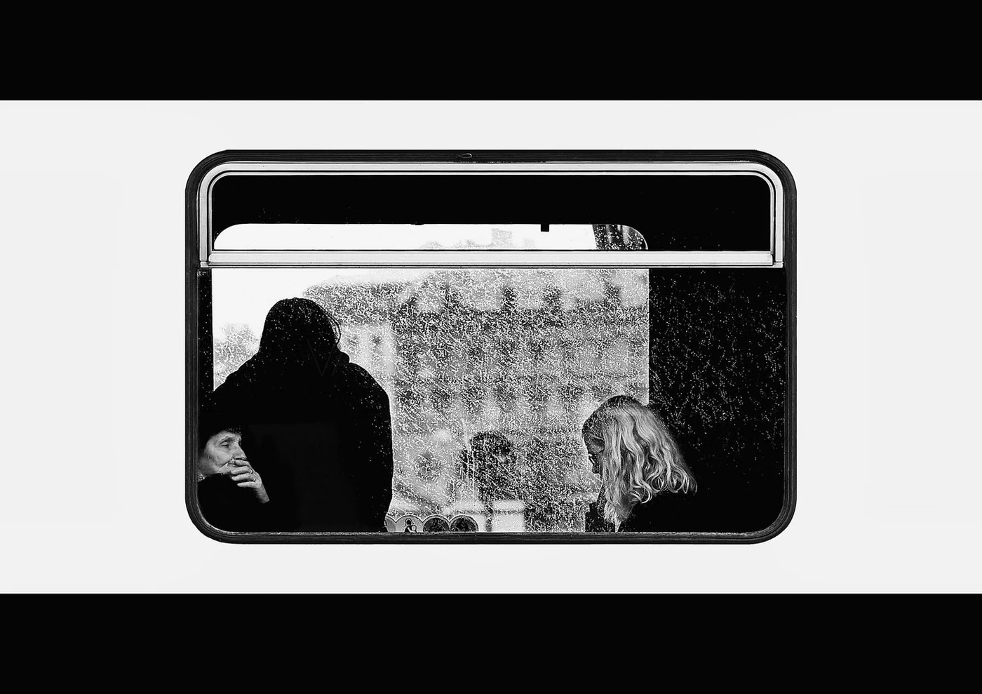 Three people inside a rain-covered bus window, two women and one man, engaging in conversation on a rainy day.