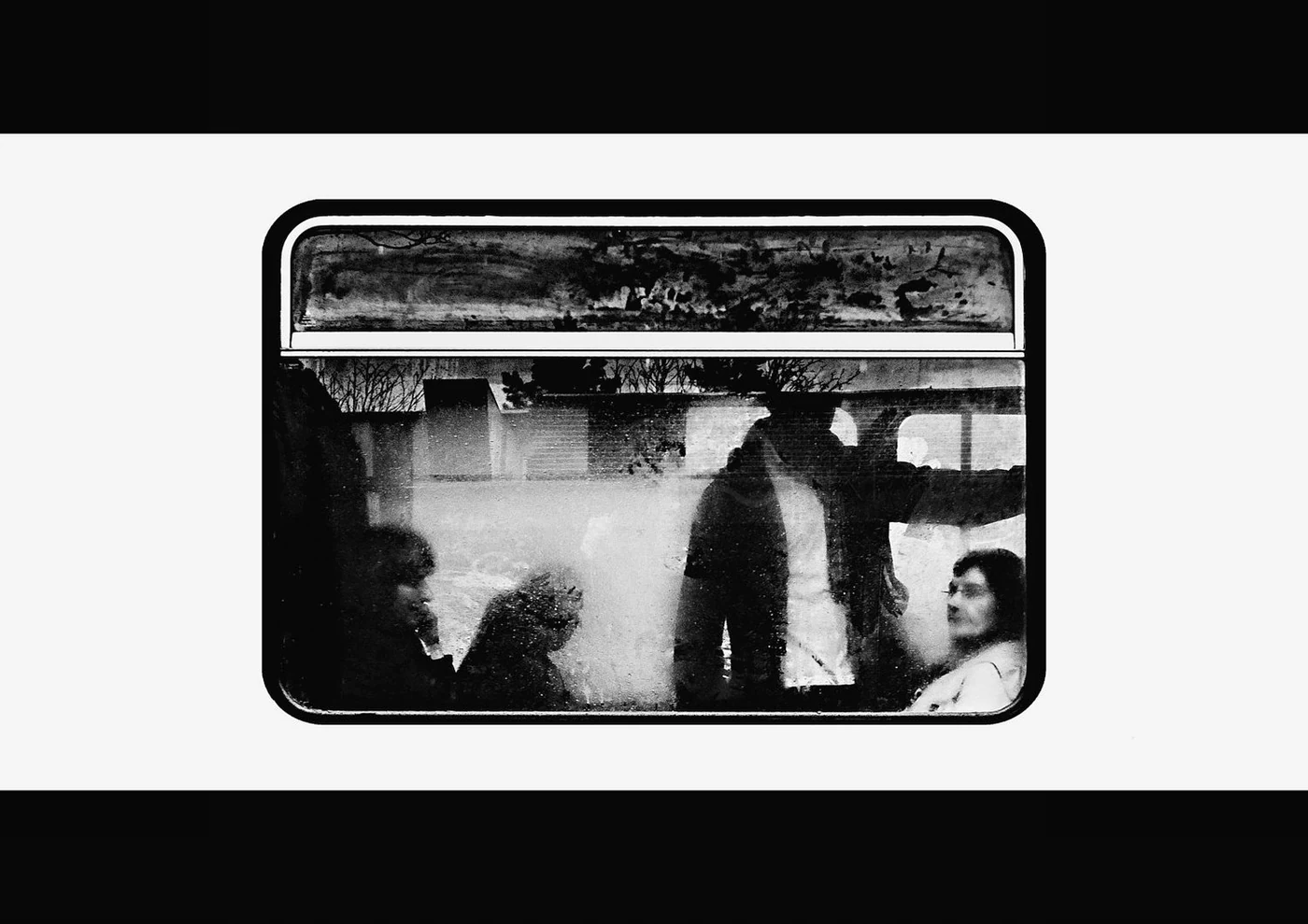 Black and white photo of four people sitting and standing inside a train, looking through the window, with trees visible outside.