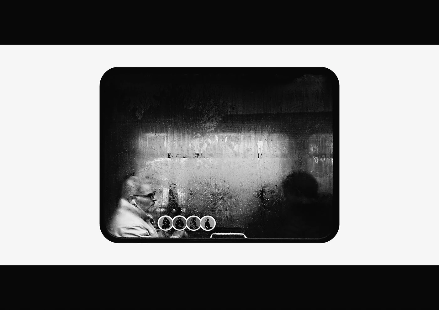 View of two people sitting on a bus during a rainy day, seen through a rain-covered window.